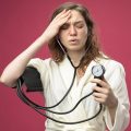 weak young ill girl with closed eyes wearing white robe measuring her own pressure with sphygmomanometer putting hand on forehead isolated on pink background weak young ill girl with closed eyes wearing white robe measuring her own pressure with sphygmomanometer putting hand on forehead isolated on pink background